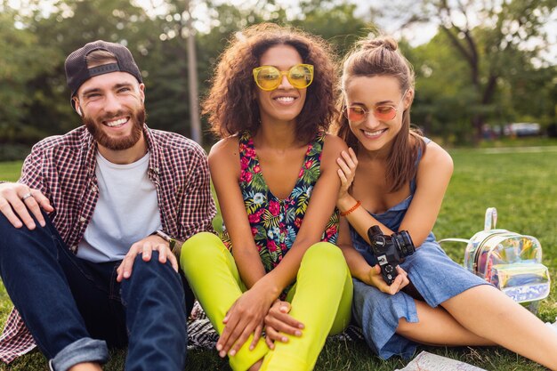 Felice giovane compagnia di amici seduti al parco, uomini e donne che si divertono insieme, viaggiano con la macchina fotografica, parlano, sorridono