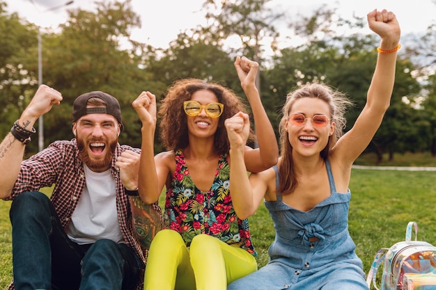 Бесплатное фото Счастливая молодая компания улыбающихся друзей, сидящих в парке на траве, мужчины и женщины, весело проводящие время вместе