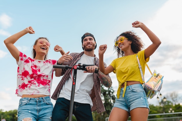 Happy young company of smiling friends dancing walking in street with electric kick scooter, man and women having fun together