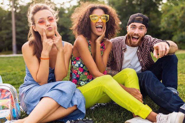 Счастливая молодая компания улыбающихся друзей, сидящих в парке на траве, мужчины и женщины, веселятся вместе, путешествуют, сумасшедшее смешное выражение лица