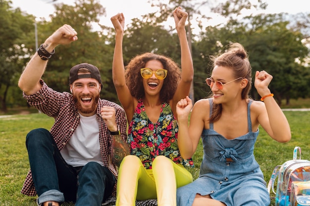 Happy young company of smiling friends sitting park on grass, man and women having fun together