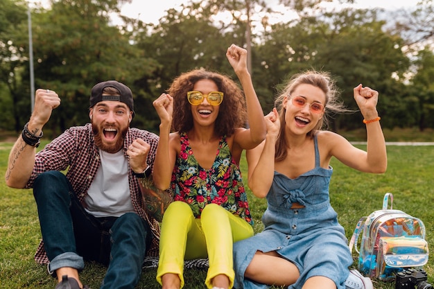 Счастливая молодая компания улыбающихся друзей, сидящих в парке на траве, мужчины и женщины, весело проводящие время вместе