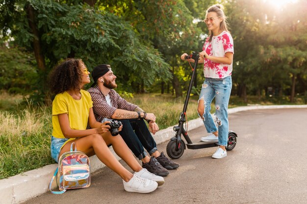 Happy young company of smiling friends sitting in park on grass with electric kick scooter, man and women having fun together