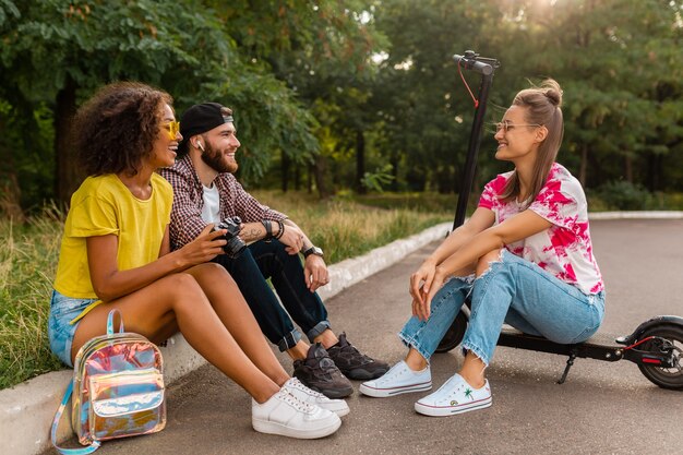 Счастливая молодая компания улыбающихся друзей, сидящих в парке на траве с электрическим самокатом, мужчины и женщины, весело проводящие время вместе