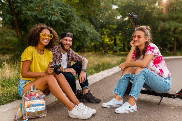 Happy young company of smiling friends sitting in park on grass with electric kick scooter, man and women having fun together