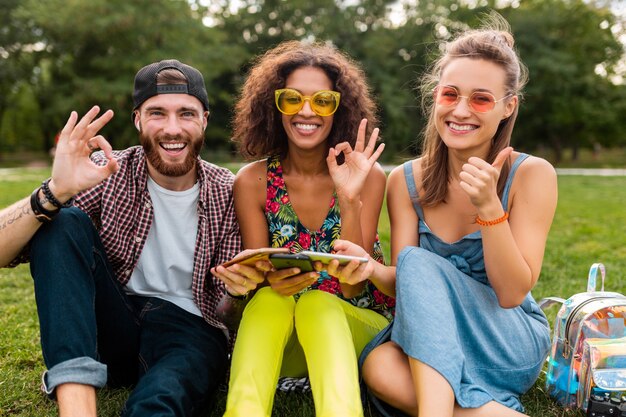 スマートフォンを使用して公園に座って、一緒に楽しんでいる笑顔の友人の幸せな若い会社カラフルな夏のスタイル、通信ワイヤレス接続デバイス、カメラで前向きに見える