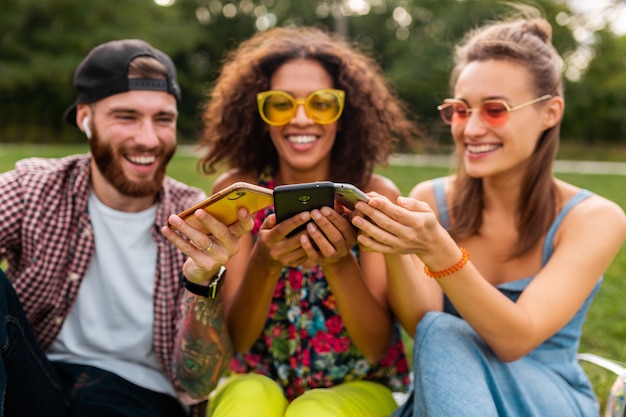 Felice giovane compagnia di amici sorridenti seduti nel parco utilizzando smartphone, uomini e donne che hanno divertimento insieme