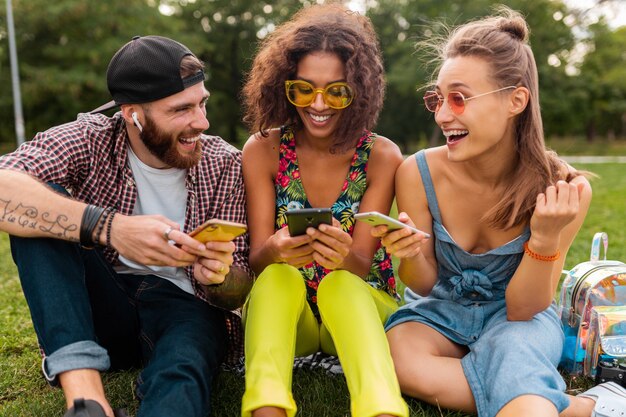 Happy young company of smiling friends sitting park using smartphones, man and women having fun together