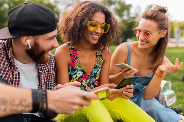 Free photo happy young company of smiling friends sitting park using smartphones, man and women having fun together