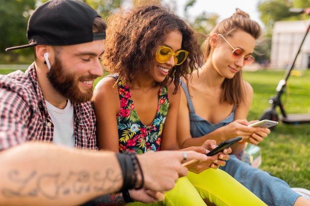 Happy young company of smiling friends sitting park using smartphones, man and women having fun together