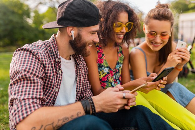 Happy young company of smiling friends sitting park using smartphones, man and women having fun together