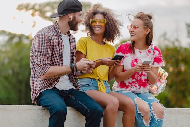 Happy young company of smiling friends sitting park using smartphones, man and women having fun