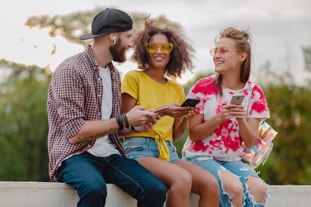 Happy young company of smiling friends sitting park using smartphones, man and women having fun