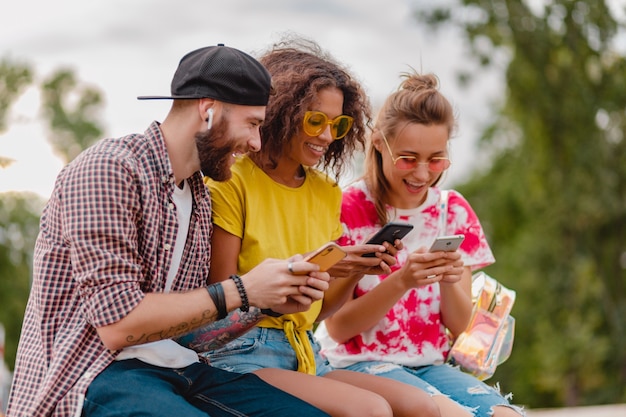 Счастливая молодая компания улыбающихся друзей, сидящих в парке с помощью смартфонов