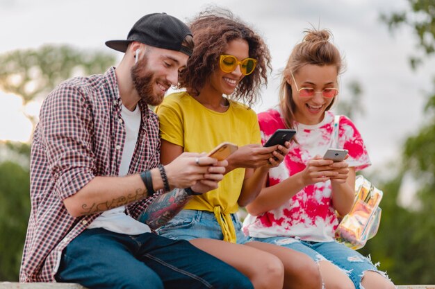 Счастливая молодая компания улыбающихся друзей, сидящих в парке с помощью смартфонов