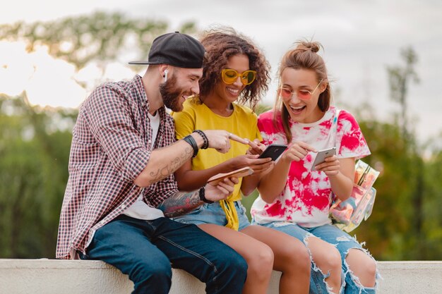 Happy young company of smiling friends sitting park using smartphones