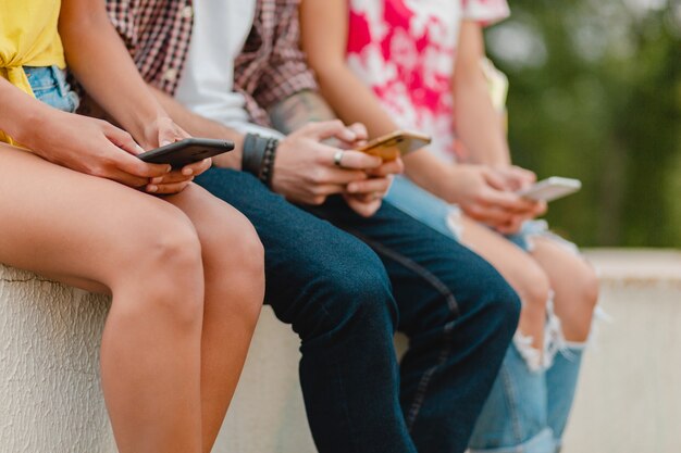 Happy young company of smiling friends sitting park using smartphones