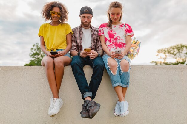 Happy young company of smiling friends sitting park using smartphones