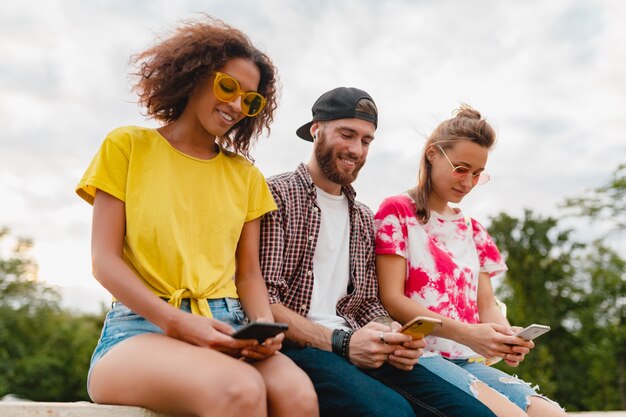 Happy young company of smiling friends sitting park using smartphones