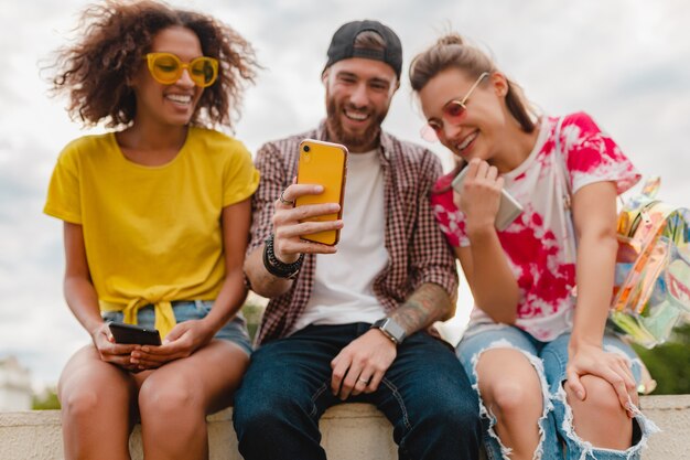 Happy young company of smiling friends sitting park using smartphones