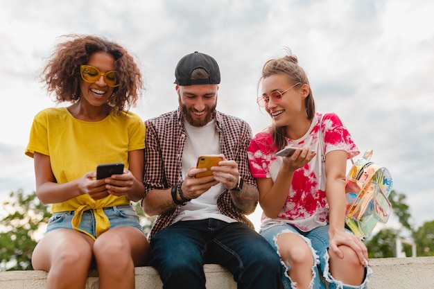 Happy young company of smiling friends sitting park using smartphones