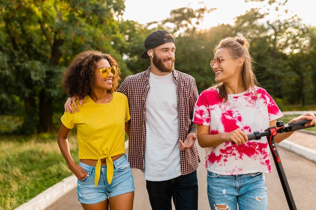 Happy young company of smiling friends walking in park with electric kick scooter, man and women having fun together