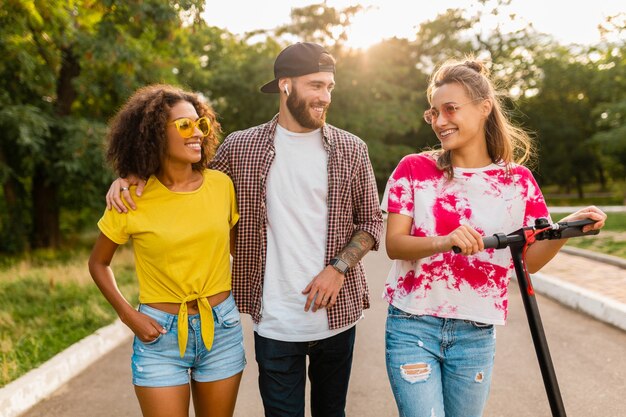 Happy young company of smiling friends walking in park with electric kick scooter, man and women having fun together