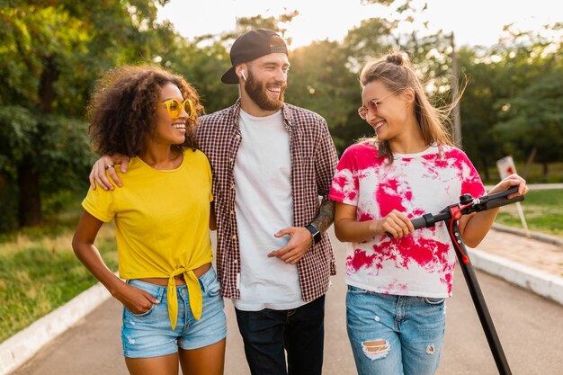 Happy young company of smiling friends walking in park with electric kick scooter, man and women having fun together