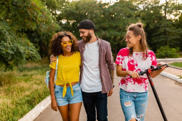 Happy young company of smiling friends walking in park with electric kick scooter, man and women having fun together