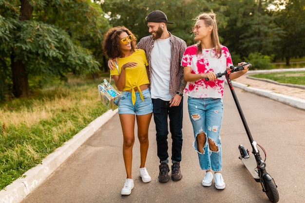 Happy young company of smiling friends walking in park with electric kick scooter, man and women having fun together