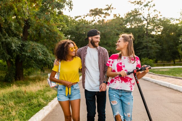 Happy young company of smiling friends walking in park with electric kick scooter, man and women having fun together