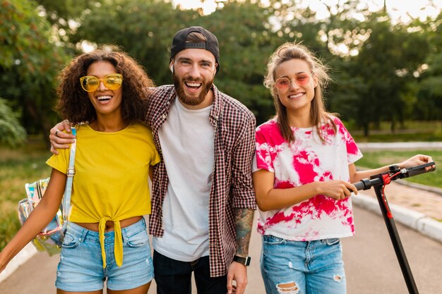 Happy young company of smiling friends walking in park with electric kick scooter, man and women having fun together