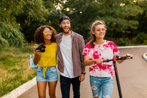 Happy young company of smiling friends walking in park with electric kick scooter, man and women having fun together