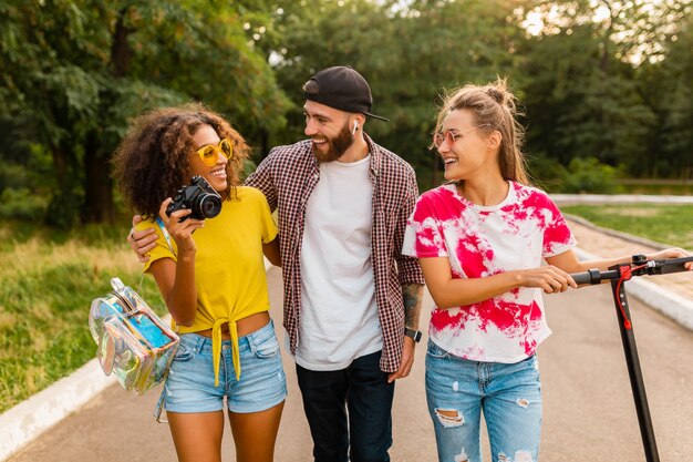 Happy young company of smiling friends walking in park with electric kick scooter, man and women having fun together