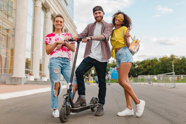 Happy young company of smiling friends walking in street with electric kick scooter, man and women having fun together