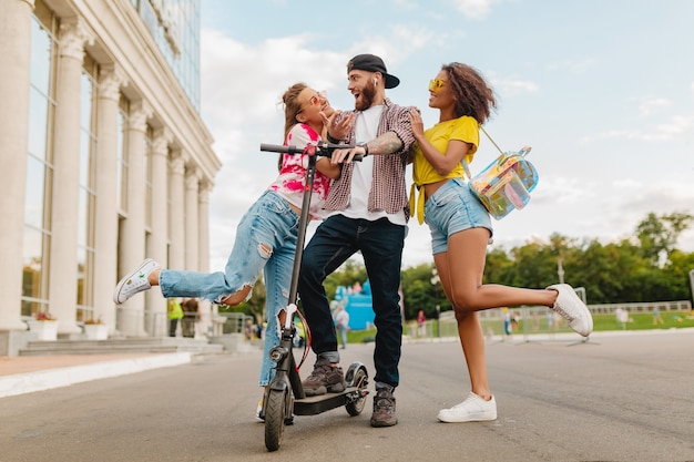 Happy young company of smiling friends walking in street with electric kick scooter, man and women having fun together