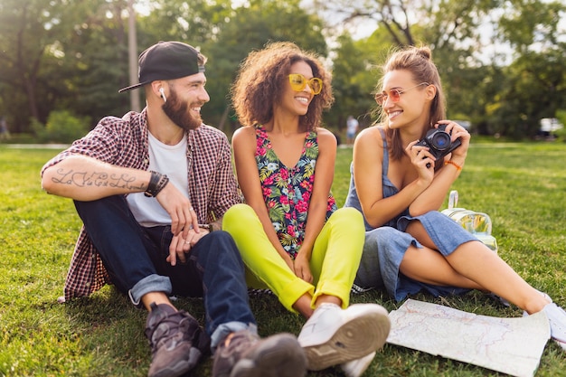 Happy young company of talking smiling friends sitting park, man and women having fun together, colorful summer hipster fashion style, traveling with camera