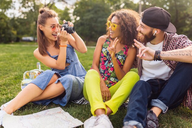 Happy young company of talking smiling friends sitting park, man and women having fun together, colorful summer hipster fashion style, traveling with camera