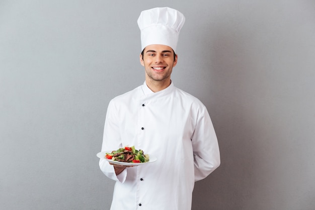 Happy young cook in uniform holding salad.