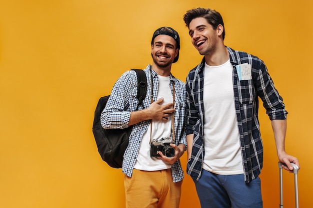 Happy young cool brunet men in white t-shirts and checkered shirts rejoice, smile and pose on orange wall. Travelers hold backpack and retro camera.