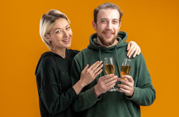 Free photo happy young couple in casual clothes mand with glasses of champagne and cheerful woman  smiling broadly celebrating valentines day standing over orange wall