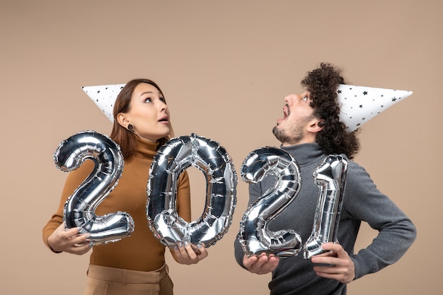 Free photo happy young couple looking above wear new year hat poses for camera girl showing and and guy with and on gray