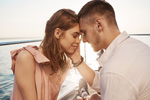 笑顔と海でのボート旅行を楽しんで愛の幸せな若いカップル。ロマンスと休暇の概念。ボーイフレンドは彼女の頬に優しく触れ、ガールフレンドは胃の中で蝶を感じる