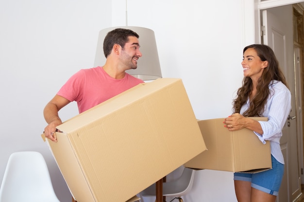 Happy young couple moving into new apartment, carrying carton boxes, looking around and smiling