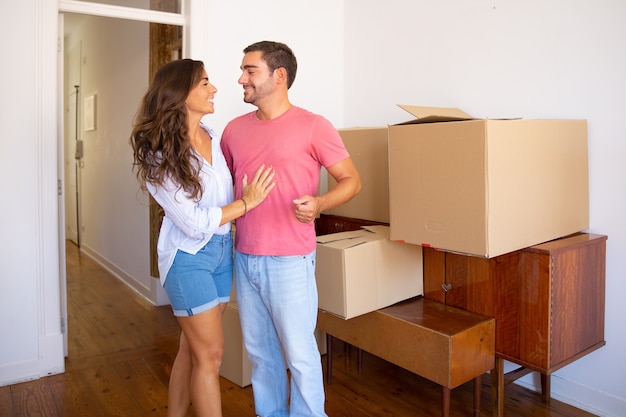 Happy young couple moving into new flat, standing near furniture and carton boxes and discussing unpacking