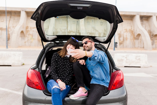 Happy young couple sitting in car trunk taking self portrait