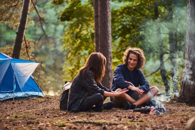 Free photo happy young couple sitting and warming near a campfire at camp in the forest. travel, tourism, and hike concept.