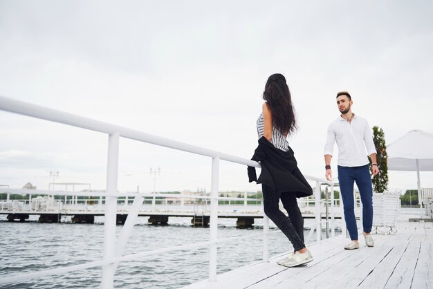 Happy young couple in stylish branded clothes, standing on the pier in the water.