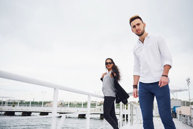 Happy young couple in stylish branded clothes, standing on the pier in the water.