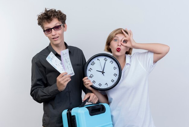 Happy young couple of tourists man holding holding air tickets and suitcase with woman holding wall clock doing ok sing standing over white wall